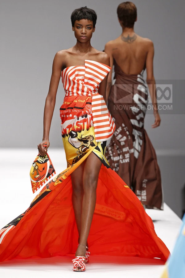 Model wearing a strapless orange and white striped dress with a graphic print and matching clutch on a runway.