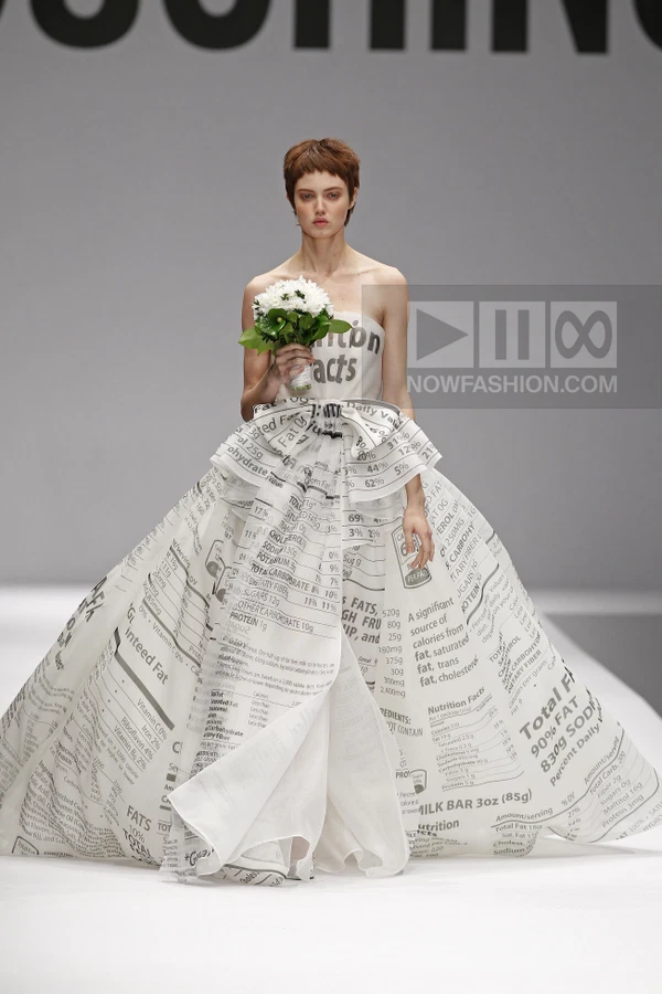 Model wearing a white strapless ball gown printed with nutrition facts text, holding a small bouquet of white flowers.