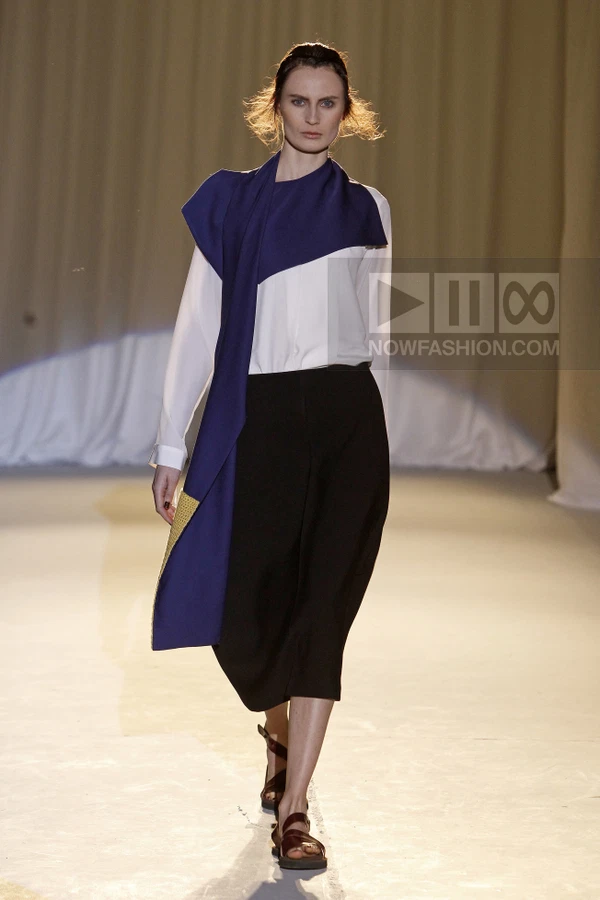Model walking in a white shirt, black skirt, and blue scarf, wearing brown flat sandals on a beige runway.