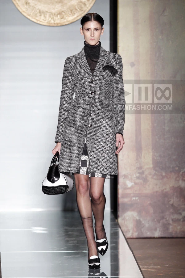 Model wearing a black and white tweed coat over a dark turtleneck and patterned skirt, carrying a black and white bag.