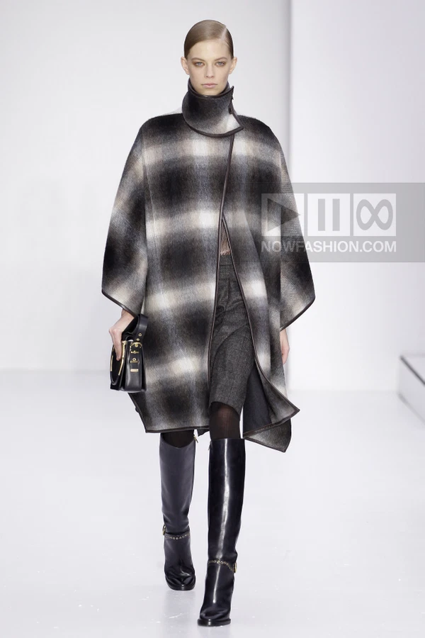 Model wearing a plaid wool cape over a dark skirt and tall black leather boots, carrying a black top-handle bag.