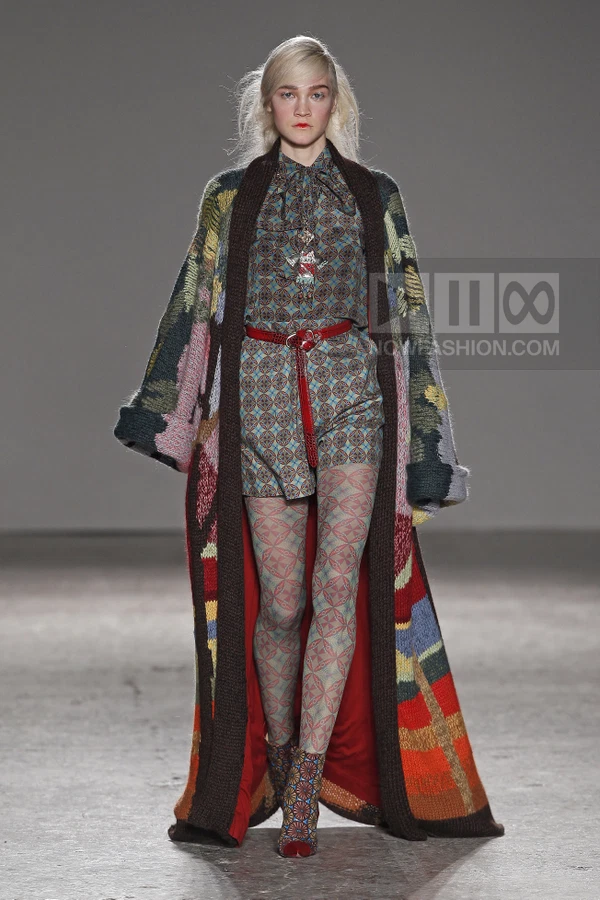 Model wearing a long, multicolored knit cardigan over a patterned blouse, shorts, and matching tights on a runway.
