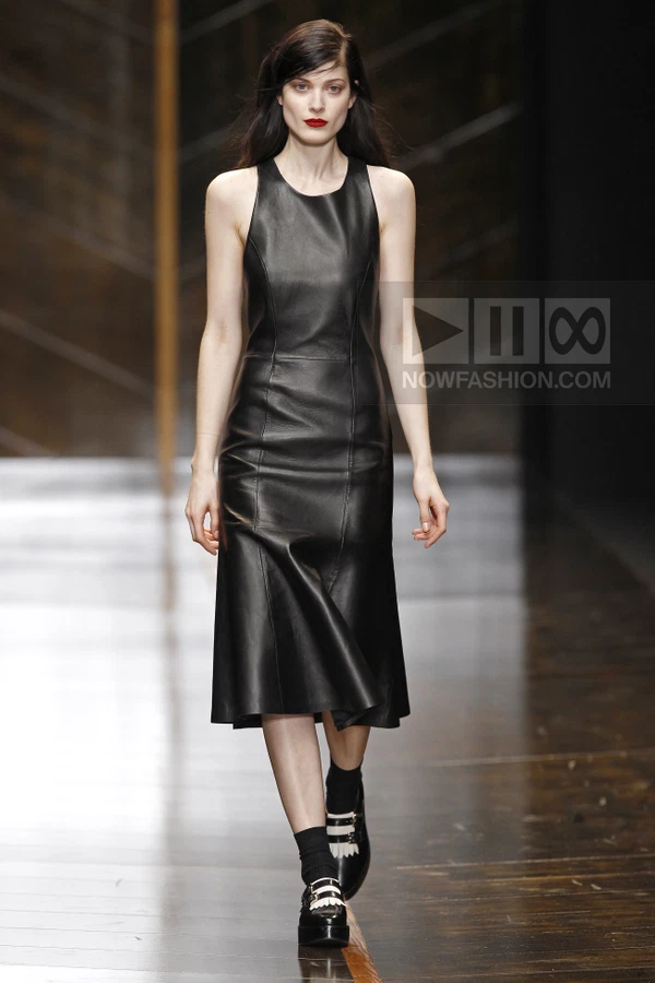 A model walks the runway in a black leather midi dress with a flared hem, paired with black socks and buckled shoes.