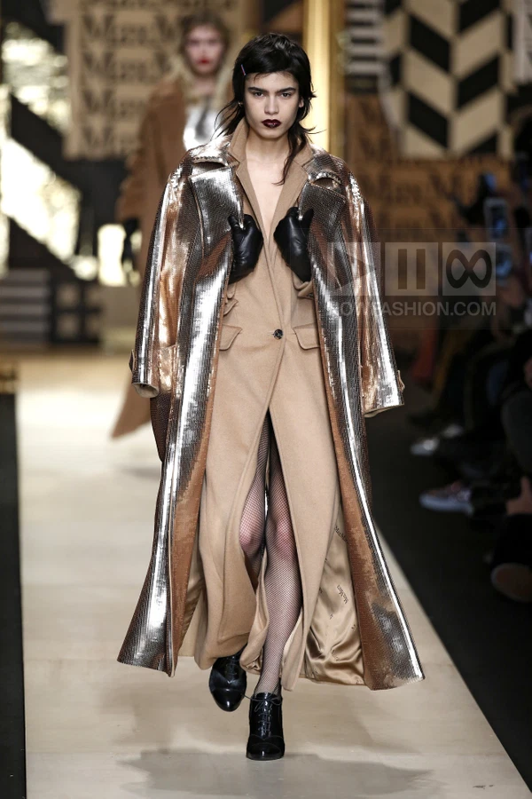 Model walking in a camel wool coat with metallic silver panels, black fishnet tights, and black lace-up shoes.