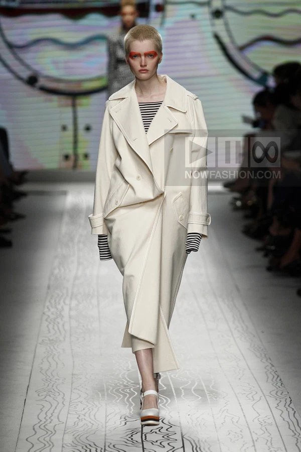 Model walking on a runway wearing an off-white double-breasted trench coat over a black and white striped top.