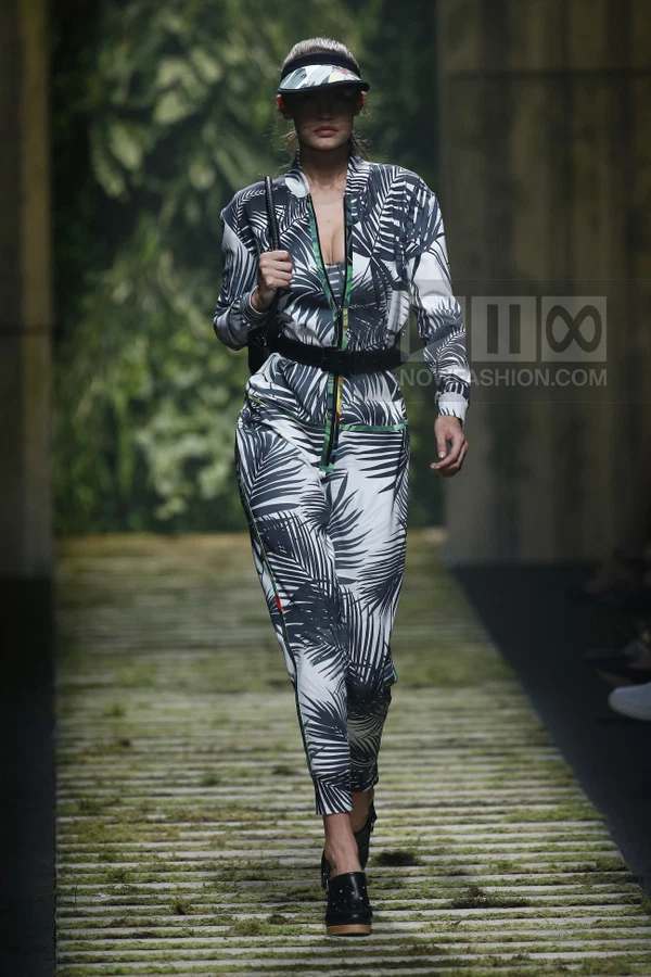 Model walking on a runway wearing a black and white palm leaf print jumpsuit, a black belt, and black mules.