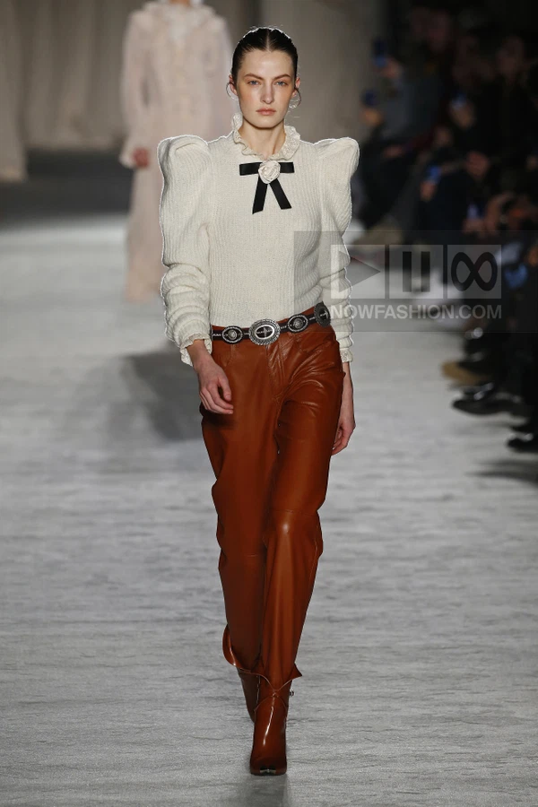 Model walking in a white knit sweater with puffed shoulders and a black bow, paired with brown leather trousers.