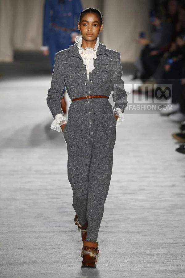 A model walks the runway in a gray wool jumpsuit with pearl buttons, a brown leather belt, and ruffled white lace trim.