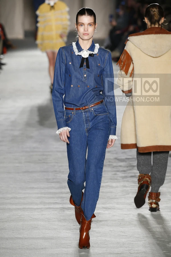 Model walking in a double denim outfit with a white lace collar and black bow, paired with brown leather ankle boots.