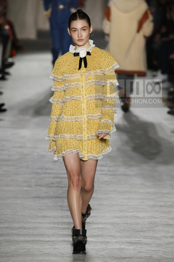 Model wearing a yellow floral mini dress with tiered lace ruffles and a black ribbon bow at the neck on a runway.