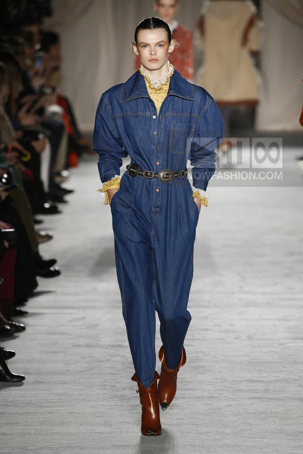 Model wearing a blue denim jumpsuit with a metal-buckled belt over a yellow floral ruffled-collar blouse on a runway.