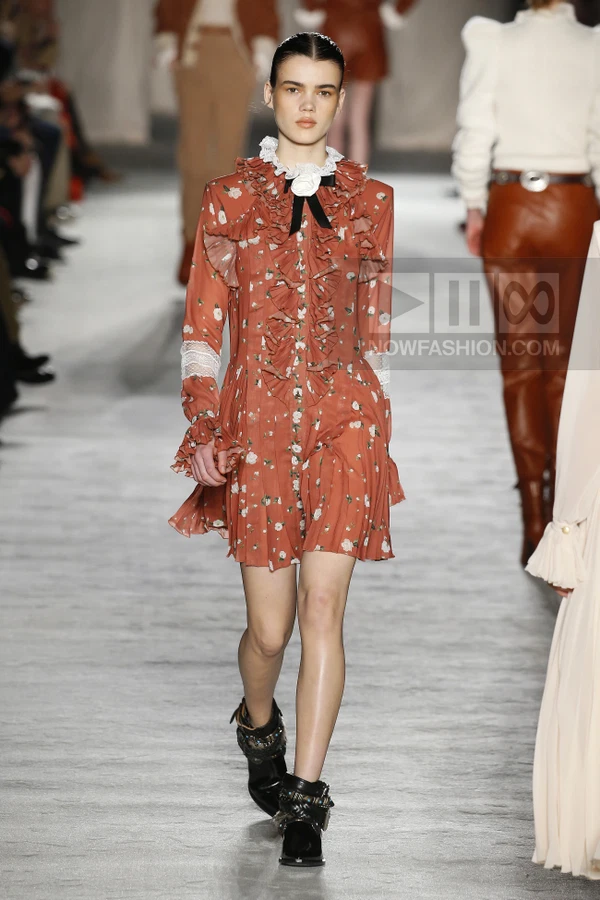 A model walks the runway in a rust-colored floral mini dress with ruffled details, lace inserts, and black ankle boots.