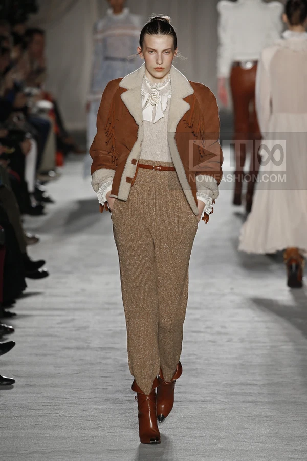 Model wearing a brown suede shearling-lined jacket with fringe, a white ruffled blouse, and brown knit trousers on runway.