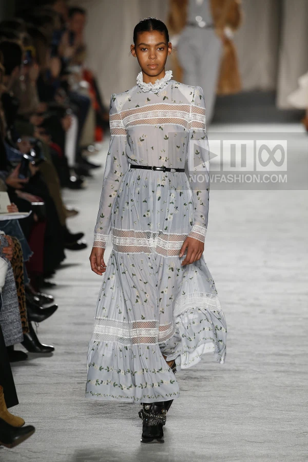 Model walking in a light blue floral midi dress with lace inserts, a high collar, and a thin black waist belt.
