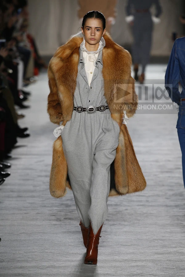 Model walking in a gray jumpsuit, brown fur coat, and brown leather boots on a runway.