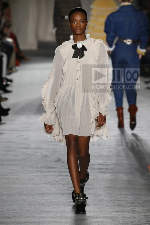 Model walking in a cream-colored chiffon mini dress with ruffles, a black bow tie, and black buckled ankle boots.