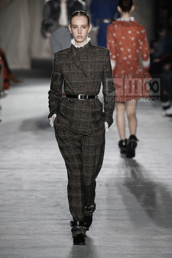 Model walking in a brown plaid wool suit with a high lace collar and black buckled belt, worn with black ankle boots.