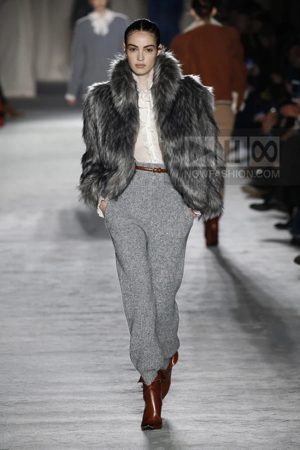 Model walking in a gray faux fur jacket, white ruffled blouse, gray knit trousers, brown belt, and brown leather boots.