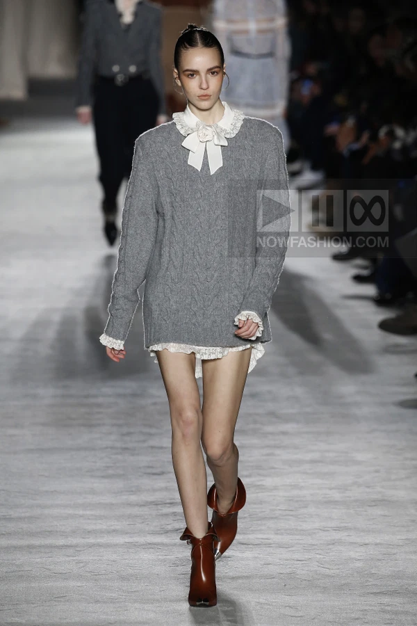 Model walking in a gray cable-knit sweater over a white lace-trimmed shirt, paired with brown leather ankle boots.