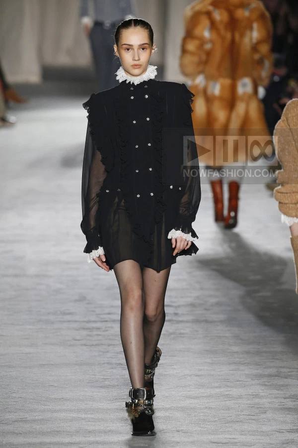A model walks the runway in a black sheer mini dress with white lace collar and cuffs, styled with dark tights and boots.