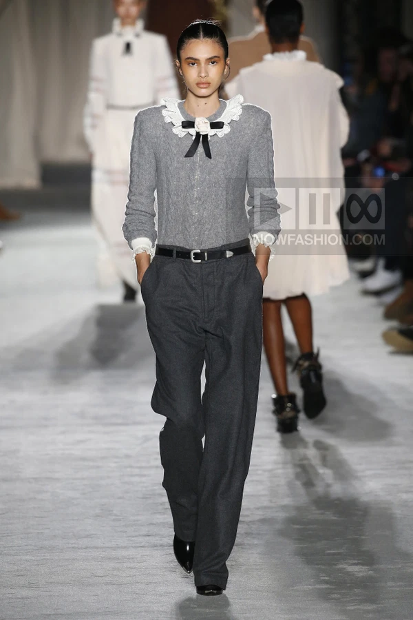 Model walking on a runway wearing a gray cable-knit sweater with a white lace collar and dark gray tailored trousers.