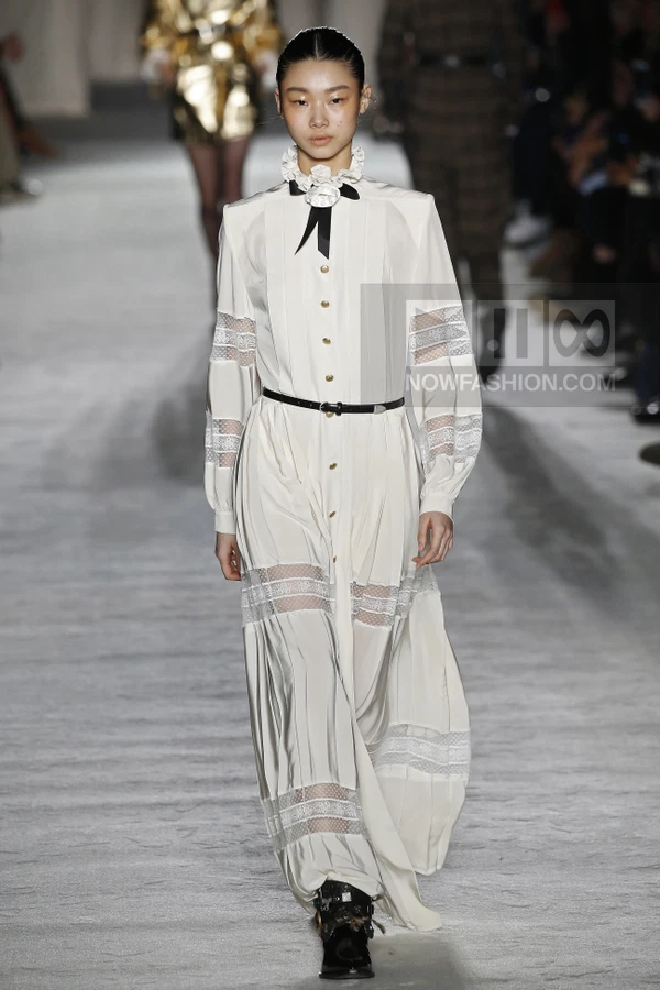 A model walks the runway wearing a white long-sleeved midi dress with lace inserts, a black ribbon tie, and a thin belt.