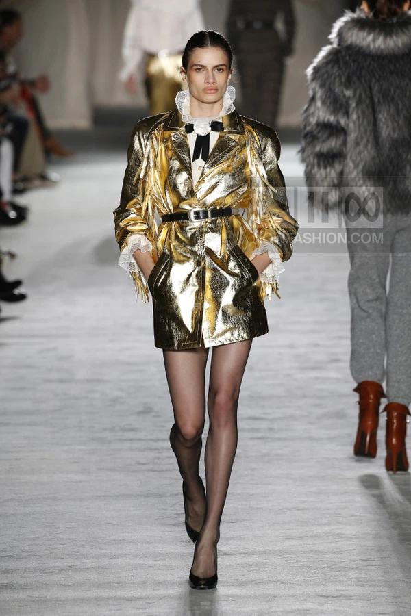 Model walks in a metallic gold jacket with fringe, white ruffled collar, and black belt over a white shirt and black tights.