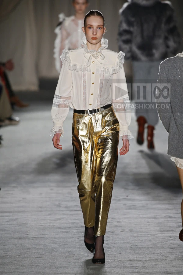 Model walking in a white ruffled blouse with lace inserts and metallic gold trousers, belted at the waist.