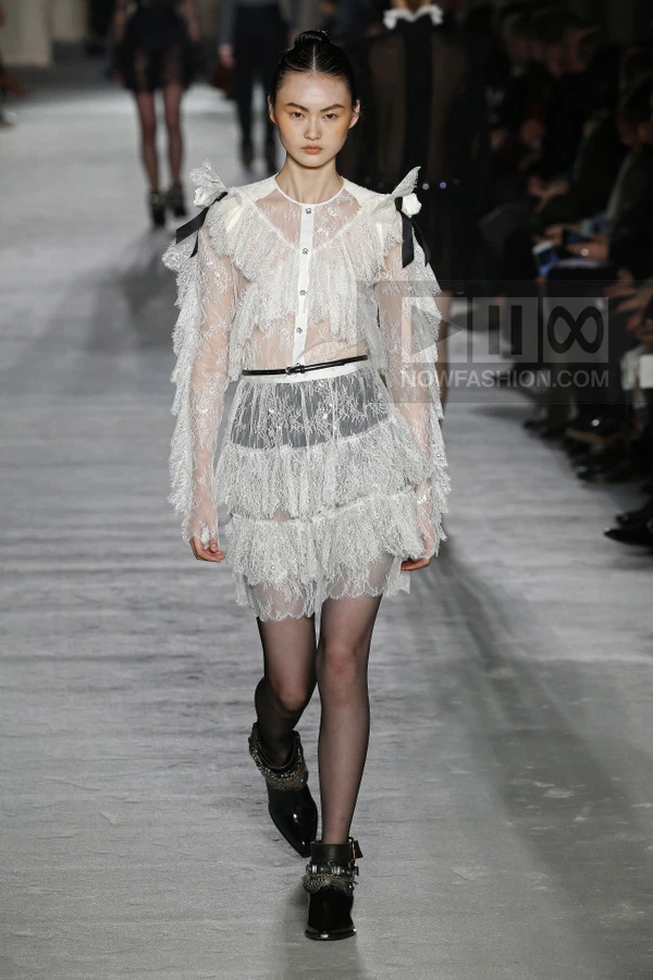 A model walks the runway wearing a white lace mini dress with ruffles, a thin black belt, and black ankle boots.