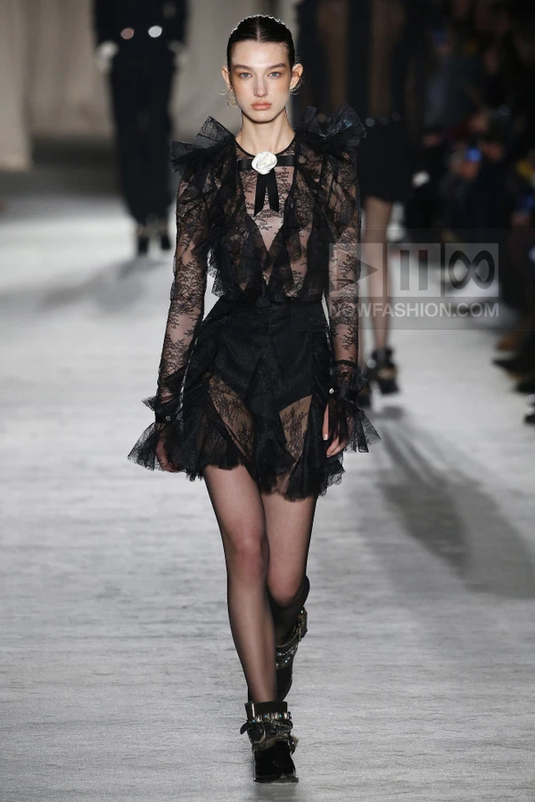 Model walking in a black lace mini dress with ruffled shoulders and a white floral brooch at the neck.