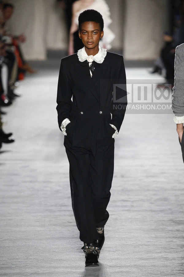 Model walking in a black double-breasted jumpsuit with a white lace-trimmed collar and cuffs on a runway.