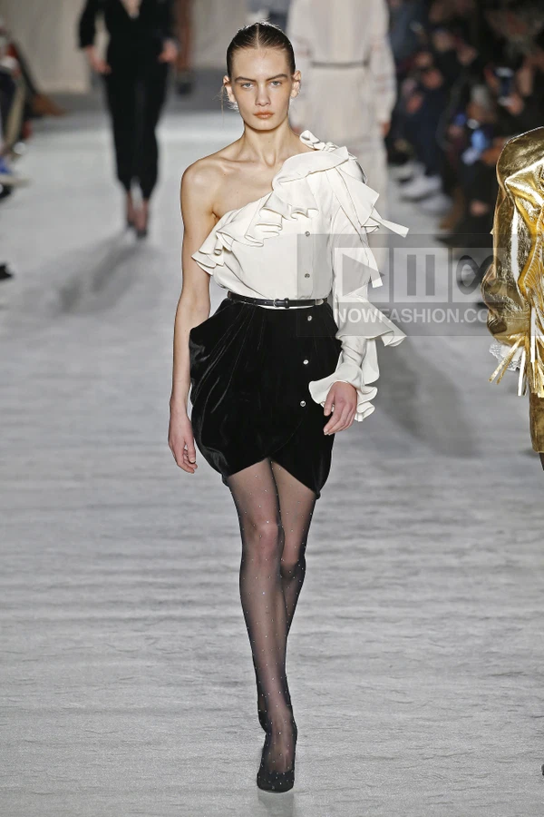Model on runway wearing white one-shoulder ruffled blouse, black velvet mini skirt, and sheer black tights.