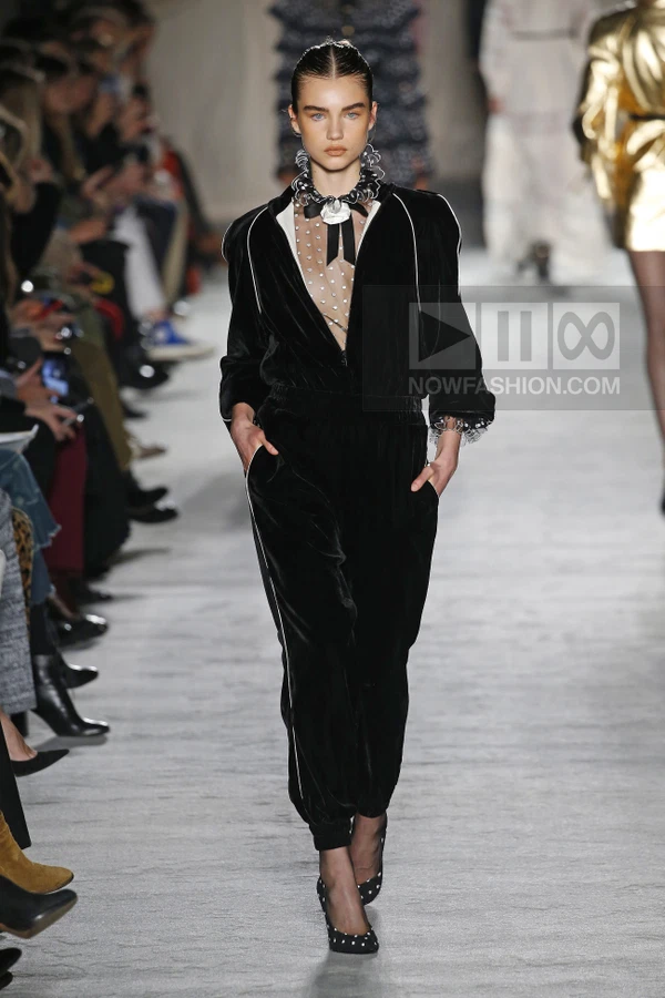 Model walking in a black velvet jumpsuit with white piping, sheer polka-dot bodice, and black pumps on a runway.
