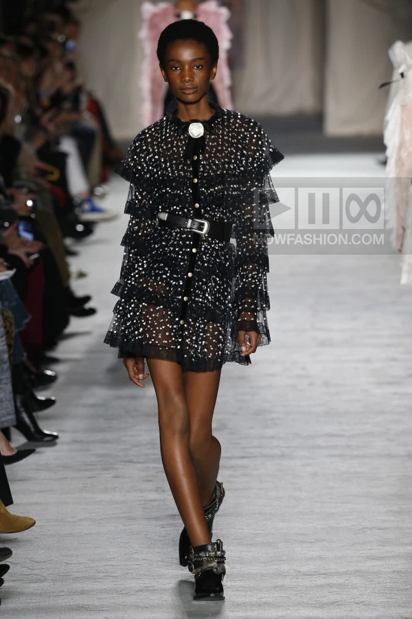 A model walks the runway in a black tiered mini dress with white polka dots, a black belt, and black ankle boots.
