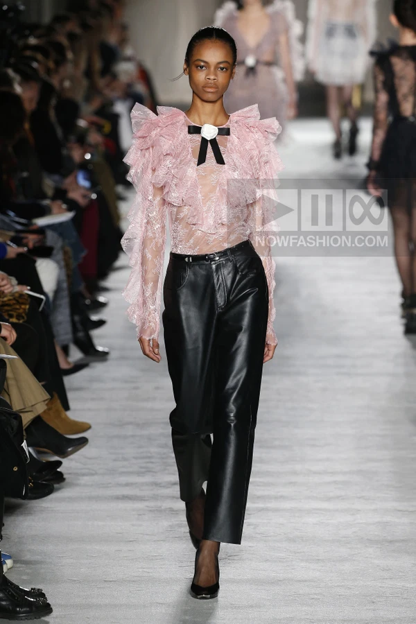 A model walks the runway wearing a pale pink lace ruffled blouse with a black bow and black leather trousers.