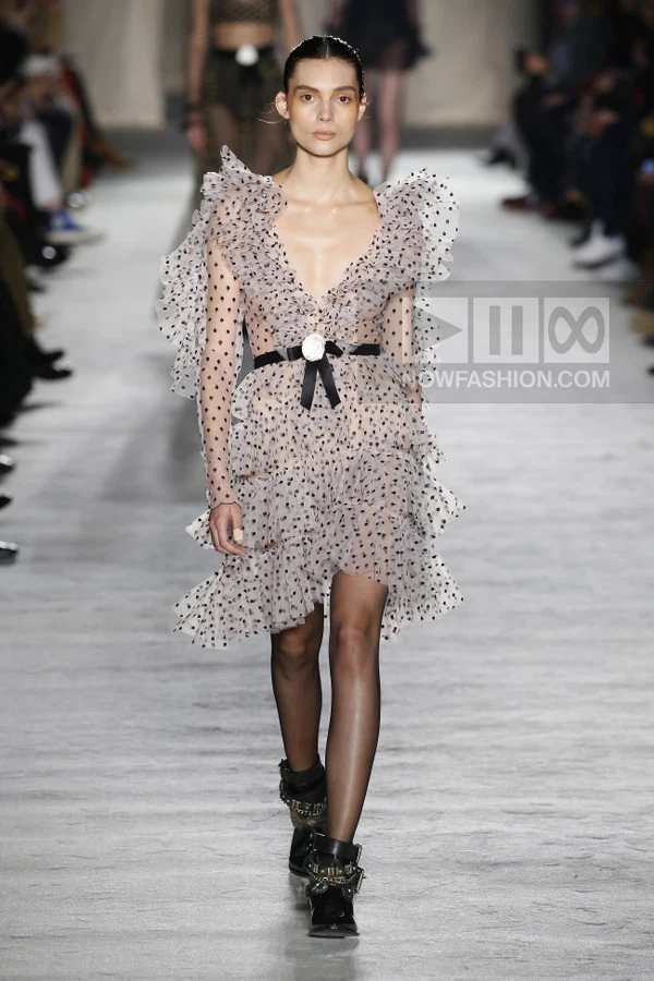 A model walks the runway in a sheer, polka-dot ruffled mini dress with a black waist belt and dark ankle boots.