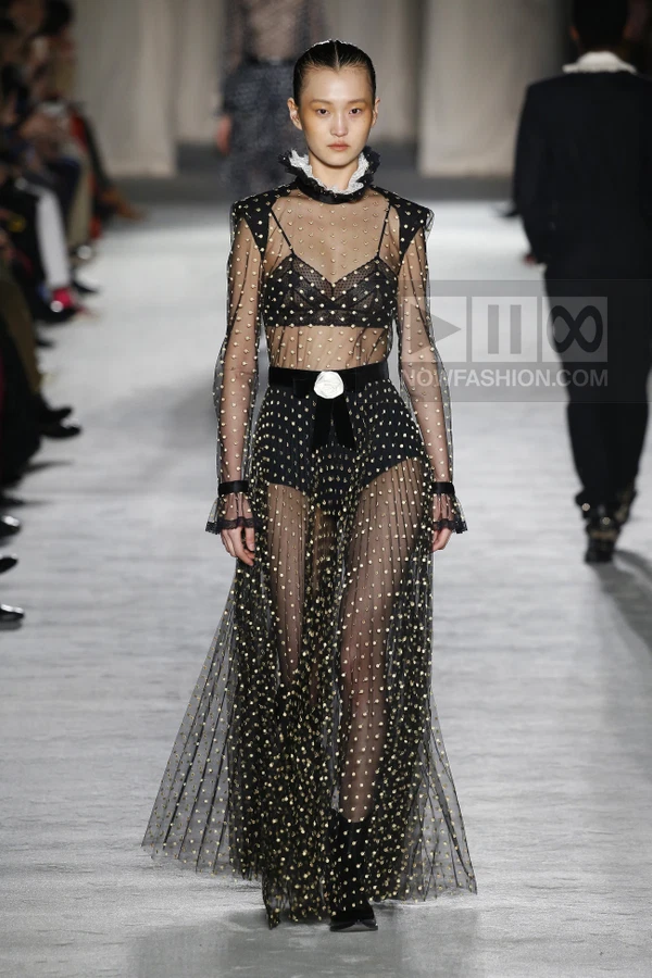 A model walks the runway in a sheer black tulle maxi dress with gold polka dots, a black bra, and a white rosette belt.