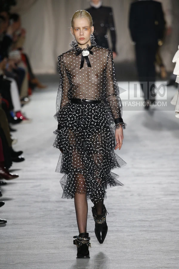 Model walking in a black sheer polka-dot tiered dress with a white rosette at the neck and black buckled ankle boots.