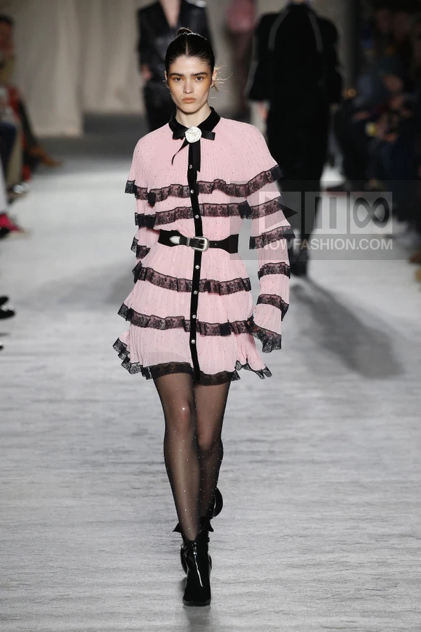 Model walks in a pink pleated mini dress with black lace trim, a black buckled waist belt, and black ankle boots.