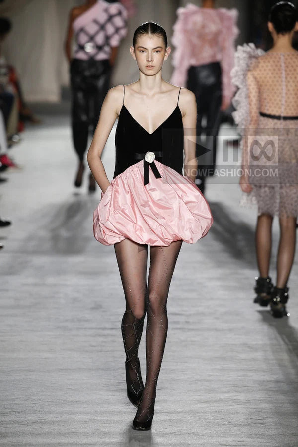 A model walks the runway in a black velvet camisole top with a pink bubble skirt and sheer patterned black tights.