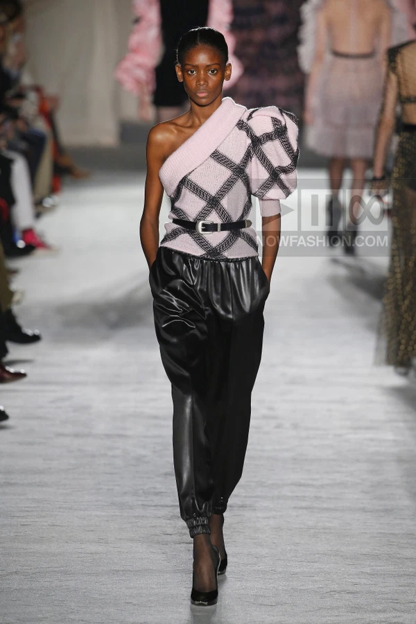 Model walking the runway wearing a pink one-shoulder knit top with black plaid, black leather joggers, and black pumps.