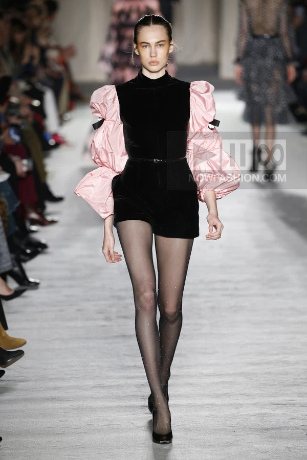 Model walking in a black velvet romper with voluminous pink satin sleeves and sheer embellished tights on a runway.