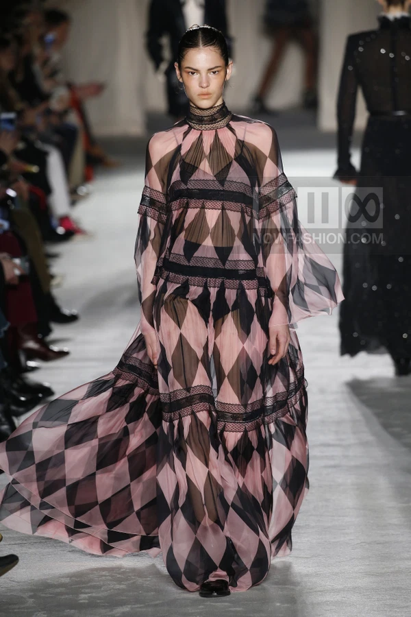 A model walks the runway in a sheer pink and black diamond-patterned maxi dress with lace trim.