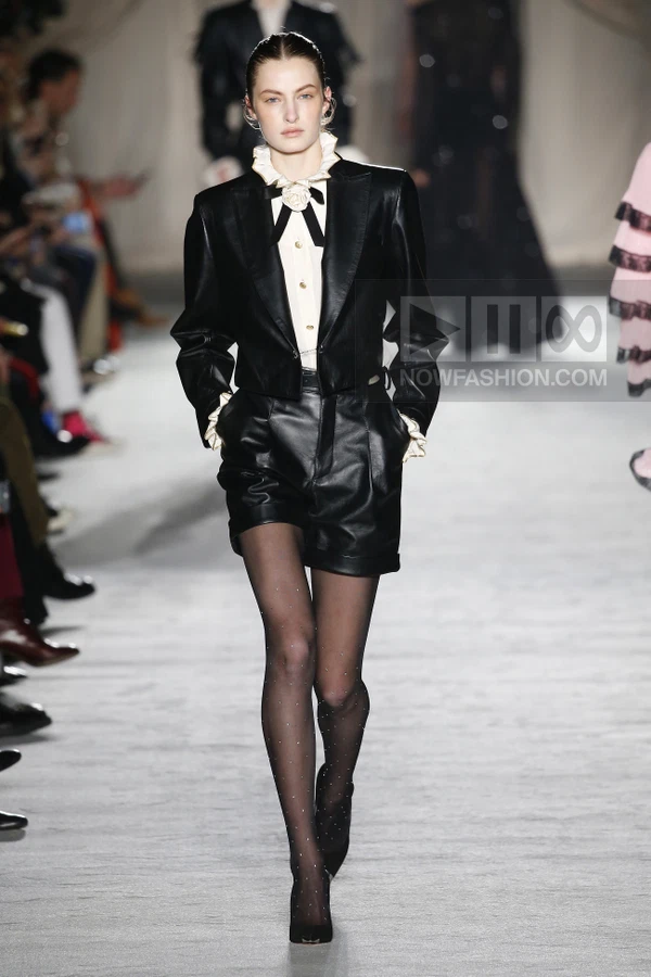 A model walks the runway wearing a black leather cropped blazer, white ruffled shirt, and black leather shorts.