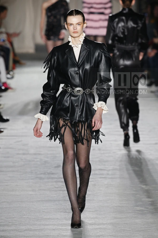 Model wearing a black leather jacket with fringe hem and ruffled collar, paired with sheer tights on a runway.