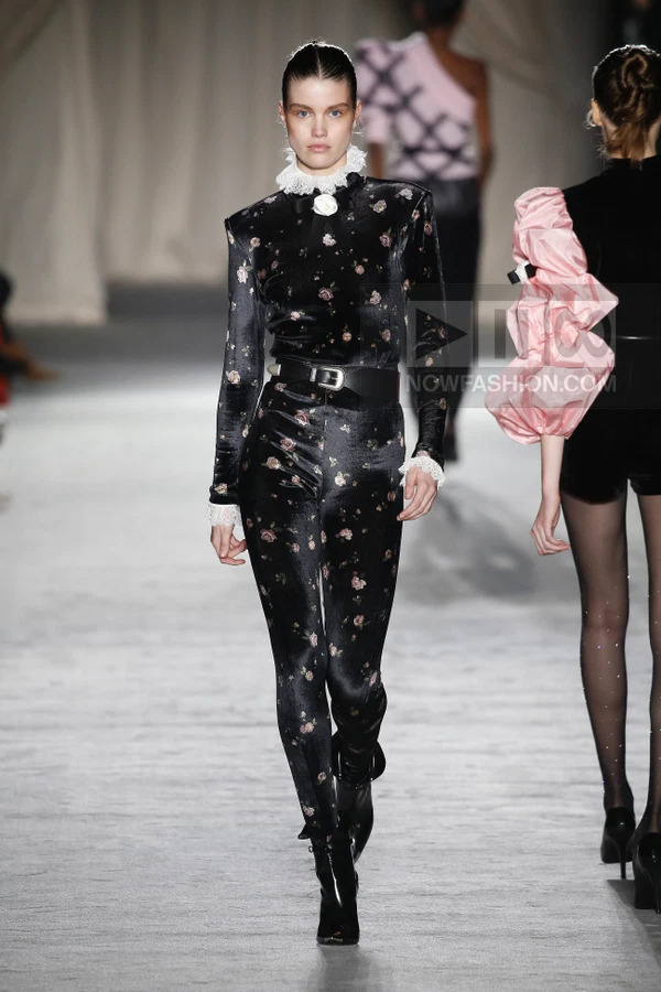 Model walking in a black velvet floral jumpsuit with a white lace collar and a black buckled waist belt.