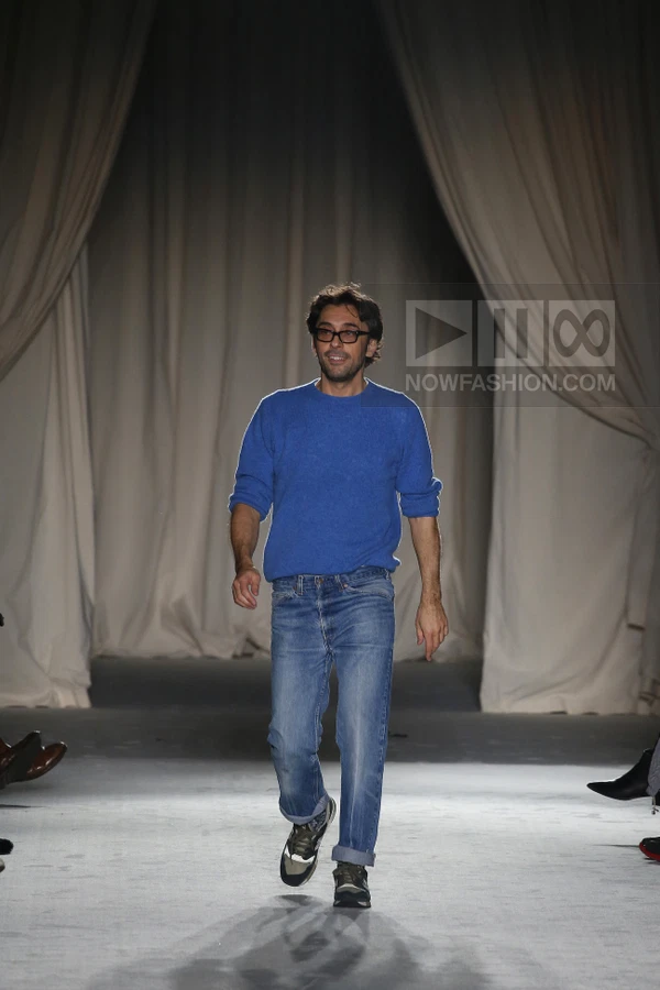 A person walking on a runway wearing a blue crew-neck sweater, blue jeans with rolled cuffs, and dark sneakers.