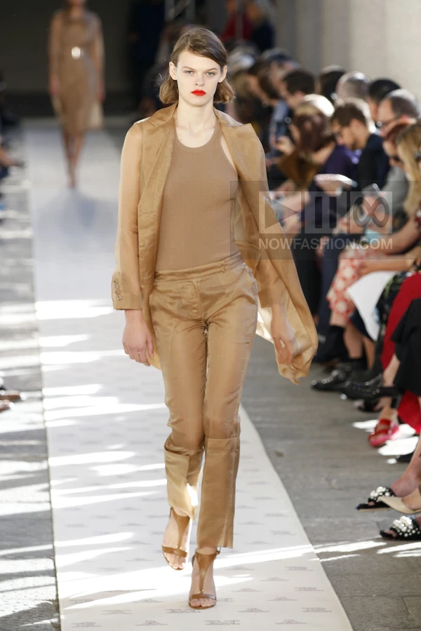 Model walking in a sheer tan coat, matching trousers, and a fitted tank top on a runway.