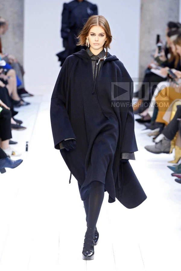 A model walks the runway in a dark navy hooded wool cape layered over a charcoal button-up shirt and black tights.