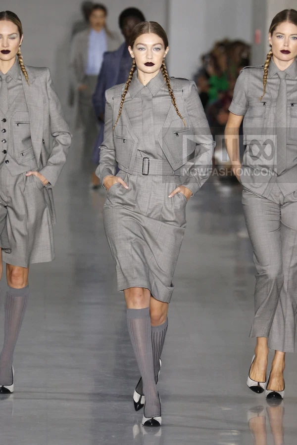 Model on runway wearing a gray wool skirt suit with a cropped jacket, belted waist, and knee-high gray socks.